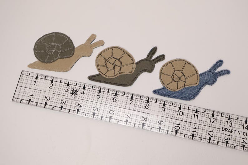 Op de afbeelding: Drie stoffen slak-applicaties met gestikte schelpdetails, geplaatst boven een doorzichtige plastic liniaal met centimeteraanduidingen. De slakken zijn beige, donkergroen en blauw van kleur, elk met een contrasterende kleur voor de slakkenhuisjes.