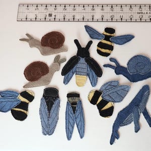 Op de afbeelding: Een verzameling stoffen patches in de vorm van insecten, slakken en een walvis, gerangschikt onder een liniaal van 38 cm. De patches hebben verschillende texturen en kleuren, waaronder spijkerstofblauw, bruin corduroy en geel-zwart gestreepte ontwerpen.