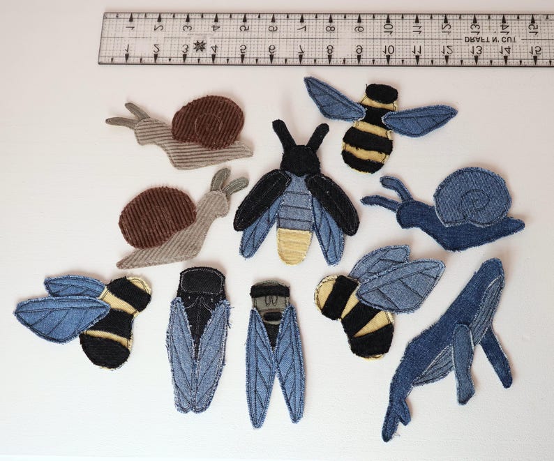 Op de afbeelding: Een verzameling stoffen patches in de vorm van insecten, slakken en een walvis, gerangschikt onder een liniaal van 38 cm. De patches hebben verschillende texturen en kleuren, waaronder spijkerstofblauw, bruin corduroy en geel-zwart gestreepte ontwerpen.
