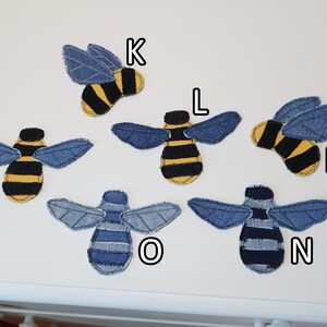 Op de afbeelding: Zes bijvormige stoffen patches gelabeld K, L, M, N, O en P. Patches K, L, M en P hebben geel en zwart gestreepte lichamen met blauwe spijkerstof vleugels. Patches N en O hebben blauw en donkerblauw gestreepte lichamen met spijkerstof vleugels.