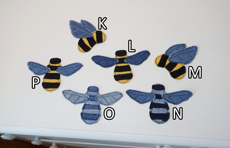 Op de afbeelding: Zes bijvormige stoffen patches gelabeld K, L, M, N, O en P. Patches K, L, M en P hebben geel en zwart gestreepte lichamen met blauwe spijkerstof vleugels. Patches N en O hebben blauw en donkerblauw gestreepte lichamen met spijkerstof vleugels.