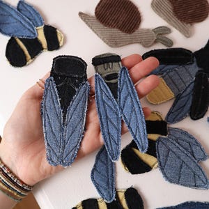 Op de afbeelding: Een collectie stoffen patches in de vorm van insecten en slakken met denim- en corduroy-texturen. De insectenpatches omvatten blauwe en zwarte cicadevormen, geel-zwart gestreepte bijen en bruine corduroy slakken met gerafelde randen.
