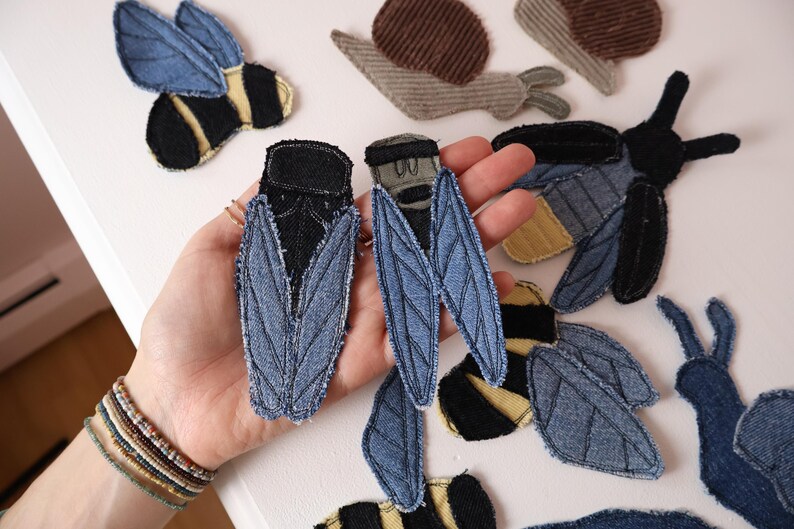 Op de afbeelding: Een collectie stoffen patches in de vorm van insecten en slakken met denim- en corduroy-texturen. De insectenpatches omvatten blauwe en zwarte cicadevormen, geel-zwart gestreepte bijen en bruine corduroy slakken met gerafelde randen.