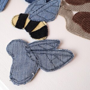 Op de afbeelding: Een verzameling stoffen patches, waaronder een bij van blauwe denim met zwarte en gele strepen, een insectvorm van blauwe denim met gestikte vleugeldetails en een slak van bruine corduroy, allemaal voorzien van zichtbaar donker stiksel.