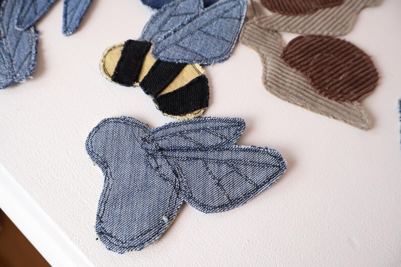 Op de afbeelding: Een verzameling stoffen patches, waaronder een bij van blauwe denim met zwarte en gele strepen, een insectvorm van blauwe denim met gestikte vleugeldetails en een slak van bruine corduroy, allemaal voorzien van zichtbaar donker stiksel.