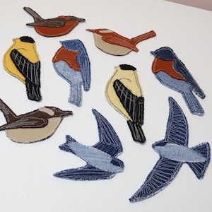 May include: A collection of nine fabric bird appliques featuring various species crafted from denim, cotton, and felt materials. The birds display diverse color palettes including shades of blue, yellow, brown, and tan, with visible stitching details.
