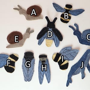 Op de afbeelding: Een verzameling van tien stoffen patches in de vorm van dieren en insecten, gelabeld van A tot J. De set bevat slakken, bijen, kevers en een walvis, gemaakt van diverse texturen denim en corduroy in blauwe, bruine, zwarte en beige tinten.