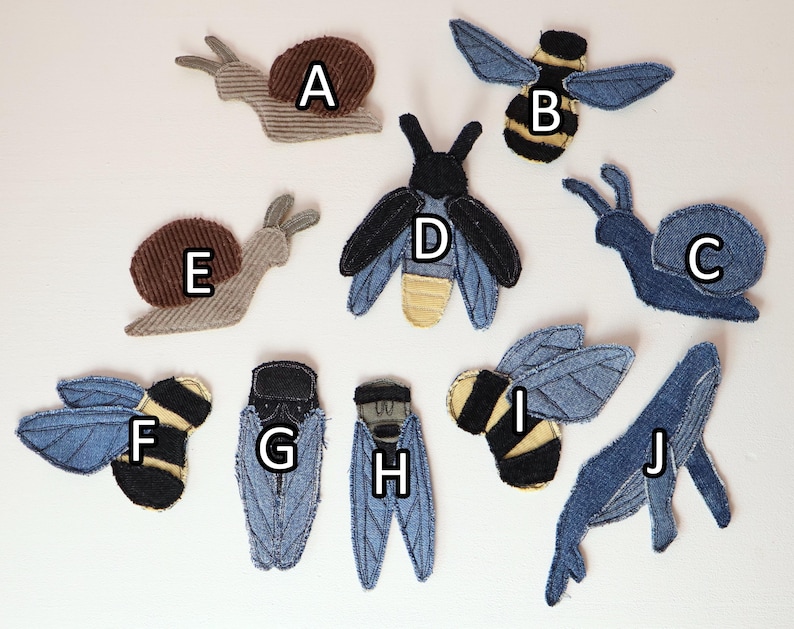 Op de afbeelding: Een verzameling van tien stoffen patches in de vorm van dieren en insecten, gelabeld van A tot J. De set bevat slakken, bijen, kevers en een walvis, gemaakt van diverse texturen denim en corduroy in blauwe, bruine, zwarte en beige tinten.