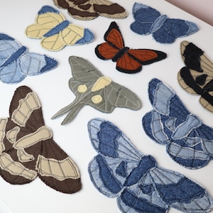 May include: A collection of ten fabric butterfly and moth appliques crafted from various shades of denim and cotton textiles. Each piece features stitched detailing, frayed edges, and distinct color patterns including blue, brown, orange, and sage green.