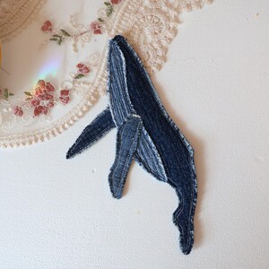 Op de afbeelding: Een walvisvormige stoffen patch gemaakt van donkerblauw denim materiaal. De patch heeft zichtbare stiksels langs de randen en het lichaam, wat zorgt voor een textuur die de vorm van een walvis met een vin en staartgedeelte nabootst.