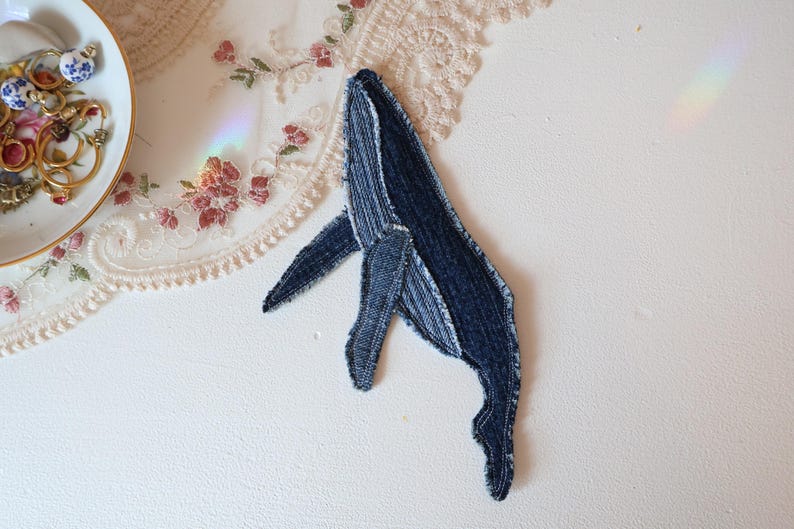Op de afbeelding: Een walvisvormige stoffen patch gemaakt van donkerblauw denim materiaal. De patch heeft zichtbare stiksels langs de randen en het lichaam, wat zorgt voor een textuur die de vorm van een walvis met een vin en staartgedeelte nabootst.
