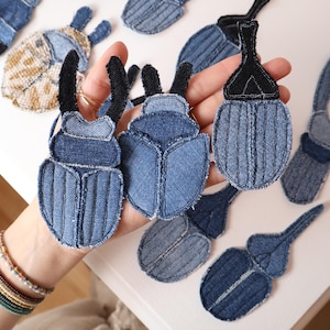 May include: A collection of beetle-shaped patches crafted from blue denim fabric. The patches feature various shades of blue denim with stitched details and some include black fabric accents for the antennae or head sections, arranged in a group.