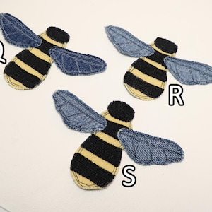 Op de afbeelding: Drie bijvormige stoffen patches met zwart-geel gestreepte lichamen en blauwe denim vleugels met gestikte aderdetails. Elke patch is gelabeld met een witte letter Q, R of S, wat duidt op verschillende variaties van het insectontwerp.