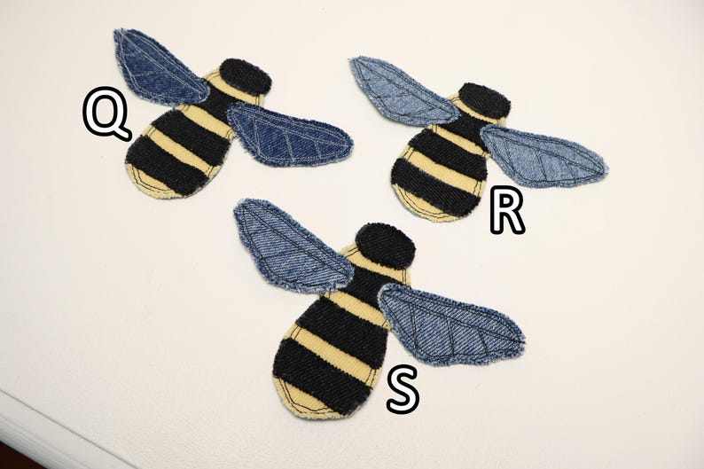Op de afbeelding: Drie bijvormige stoffen patches met zwart-geel gestreepte lichamen en blauwe denim vleugels met gestikte aderdetails. Elke patch is gelabeld met een witte letter Q, R of S, wat duidt op verschillende variaties van het insectontwerp.