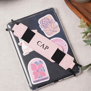 May include: A rectangular tablet case with a clear back featuring multiple decorative stickers, including one labeled FLOWER MARKET. A light pink elastic hand strap with the word CAP printed in black is attached across the center of the case.