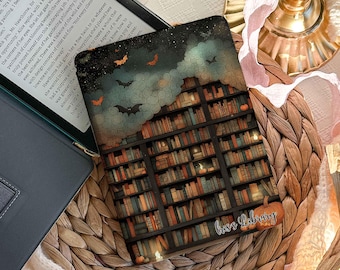 Könnte beinhalten: Eine dekorative Kindle-Hülle mit einem Bücherregal-Design, Fledermäusen und einem Kürbis. Die Hülle trägt den Text "boss library" und liegt auf einem gewebten Platzset mit einem Buch und anderen dekorativen Gegenständen.