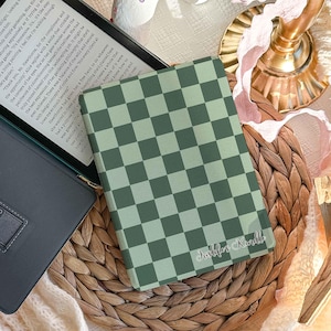 Peut inclure: Une housse de Kindle à carreaux verts et blancs repose sur un tapis tissé à côté d'une tablette. La housse porte l'inscription "Justlifes Kindle" en cursive. Une couverture douce de couleur crème et des objets décoratifs sont en arrière-plan.