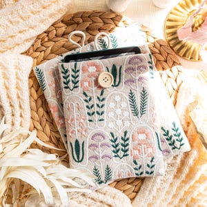 May include: A beige tablet case with a floral embroidery design. The case has a button closure and is made of a soft, textured fabric.