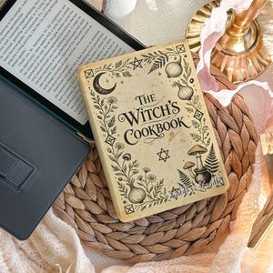 Peut inclure: Un autocollant rectangulaire pour liseuse avec un fond crème et des illustrations botaniques noires, incluant des champignons, des oignons et un croissant de lune. Le texte indique THE WITCH'S COOKBOOK avec une petite étoile en dessous.