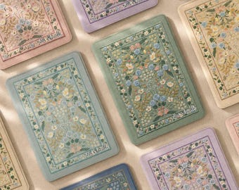 May include: A collection of rectangular fabric covers featuring intricate floral embroidery in pastel shades of pink, blue, green, and yellow. Each cover displays a central botanical pattern framed by a decorative border and the text 'has & kindle'.