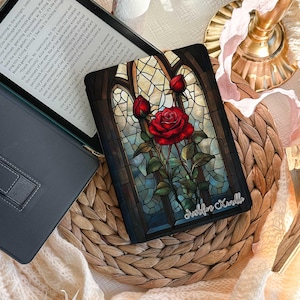 Könnte beinhalten: Eine dekorative E-Reader-Hülle mit einem Buntglas-Rosen-Design. Die schwarze Hülle zeigt eine rote Rose und grüne Blätter. Der Text "Deslilos Korallo" ist sichtbar. Sie liegt auf einer gewebten Oberfläche.