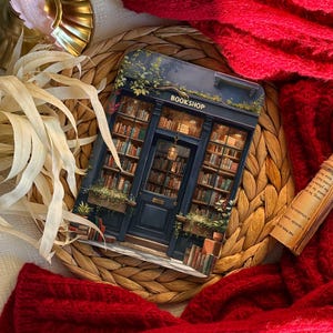May include: A decorative card featuring a detailed illustration of a bookstore facade with the text "BOOKSHOP". The card is surrounded by books, plants, and a dark blue door. The card is on a woven mat, with a red knitted scarf.