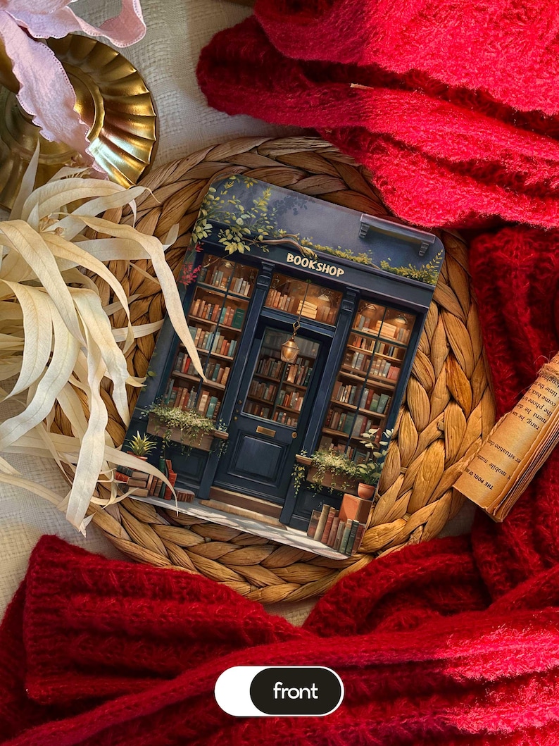 May include: A decorative card featuring a detailed illustration of a bookstore facade with the text "BOOKSHOP". The card is surrounded by books, plants, and a dark blue door. The card is on a woven mat, with a red knitted scarf.