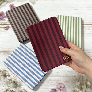 Può includere: Quattro custodie rettangolari per e-reader con strisce verticali marroni, bordeaux, verde chiaro e blu. La custodia bordeaux è tenuta in primo piano e mostra le iniziali dorate HB nell'angolo in basso a destra.