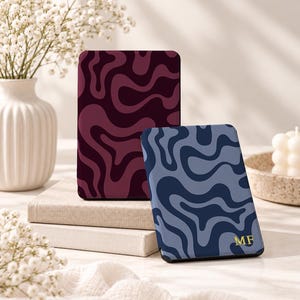 May include: Two rectangular e-reader cases featuring wavy, abstract patterns. One case is dark burgundy and maroon, while the other is navy and light blue with the gold-colored initials MF printed in the bottom right corner.