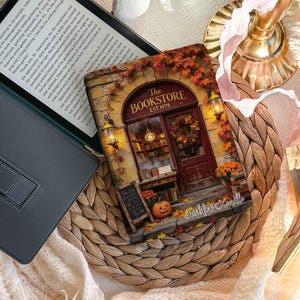 May include: A black e-reader case featuring a printed illustration of a bookstore entrance with a red door, autumn leaves, a pumpkin, and a sign that reads The Bookstore Est. 1978. The case is positioned on a round woven mat next to an open e-reader.