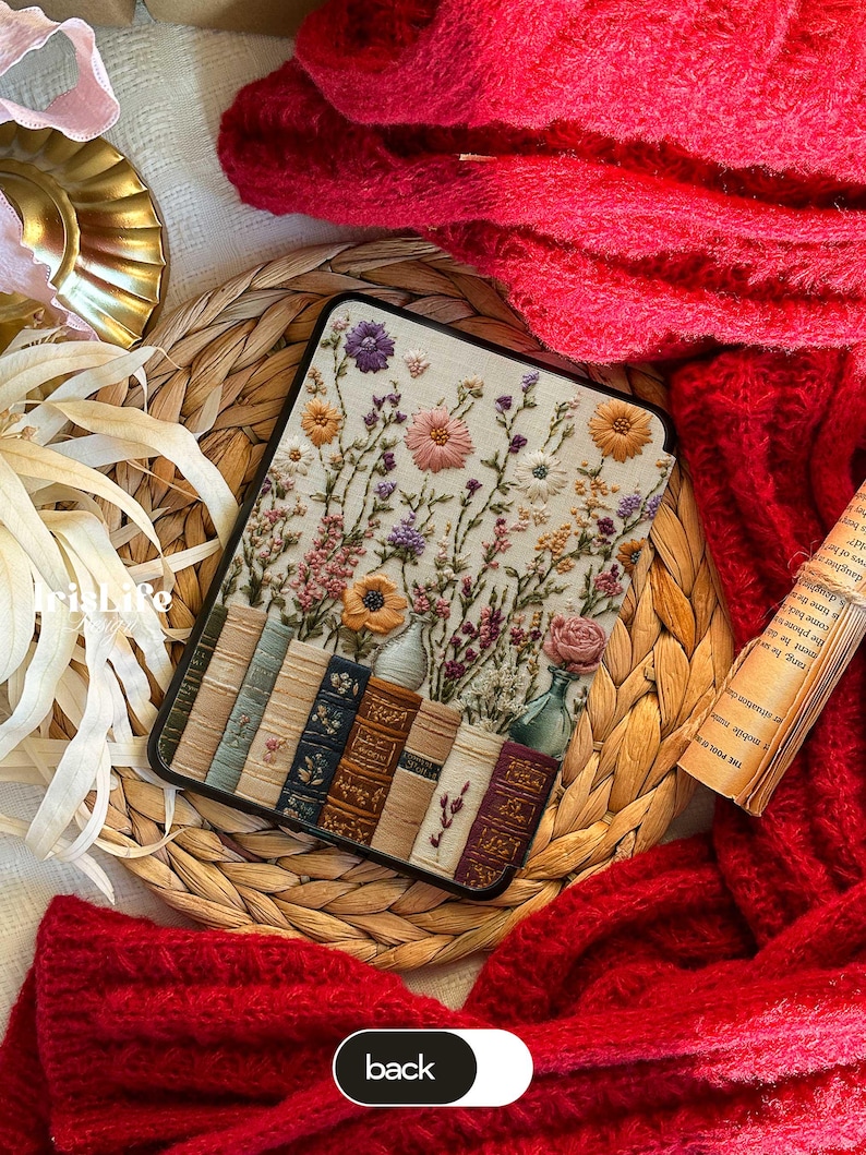 May include: A white fabric phone case with an embroidered design of flowers and books. The flowers are in shades of pink, purple, and yellow. The books are brown and have gold lettering. The case is surrounded by a woven wicker mat and a red fuzzy blanket.