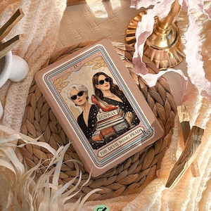 Może przedstawiać: Prostokątne etui na telefon z ilustracją dwóch kobiet w okularach przeciwsłonecznych, z napisem "RUNWAY" i "THE DEVIL WEARS PRADA". Etui jest w kolorze jasnobrązowym z ozdobną obwódką. U góry znajduje się napis "Spersonalizuj swoje etui swoim imieniem".
