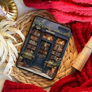May include: A phone case featuring a detailed illustration of a bookstore facade with the word "BOOKSHOP" above the entrance. The case is set on a woven mat, with a red knitted scarf and a book in the background.