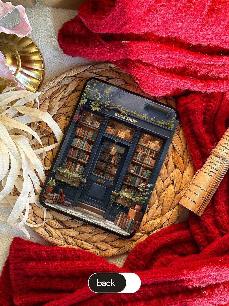 May include: A phone case featuring a detailed illustration of a bookstore facade with the word "BOOKSHOP" above the entrance. The case is set on a woven mat, with a red knitted scarf and a book in the background.