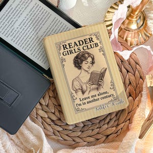 Może przedstawiać: Beżowe etui na czytnik Kindle w pionowe paski z ilustracją w stylu vintage przedstawiającą kobietę czytającą książkę. Tekst głosi "READER GIRLS CLUB" oraz "I'm in another century." Etui posiada ozdobne obramowania w formie zawijasów.