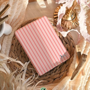 May include: A pink case with vertical stripes and the name "Isir" in cursive. The text "Personalise your case with your name" is at the top. The text "Sophia - Font #1" is at the bottom. The case is on a woven mat.