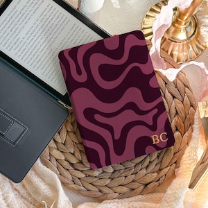 May include: A dark purple and magenta e-reader case featuring a wavy, abstract pattern and the gold-colored initials BC embossed in the bottom right corner. The case is shown closed, displaying the full front cover design with its fluid, organic shapes.