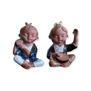May include: Two ceramic figurines of seated children, each with closed eyes. One wears a blue and white outfit and holds a fan. The other wears a black and white outfit and holds a musical instrument. Both have brown skin.