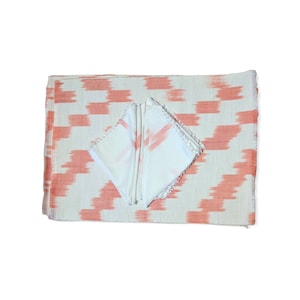 May include: A white and coral-colored table runner and matching napkins. The runner has a geometric pattern, and the napkins are folded and have a coral-colored design. The edges of the runner are fringed.