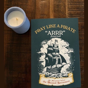 May include: A teal blue journal with the title "Pray Like A Pirate "ARRR" Journal" in gold lettering. The cover features a black and white illustration of a pirate ship sailing on a stormy sea. The text below the illustration reads "An Adventure before the Blessed Sacrament."