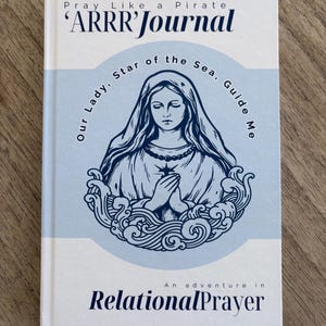 May include: A white book with a blue illustration of the Virgin Mary on the cover. The book title is "ARRR' Journal" and the subtitle is "Our Lady, Star of the Sea, Guide Me. An adventure in Relational Prayer."