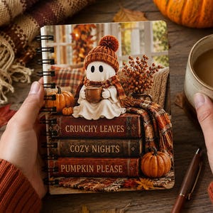 May include: A spiral-bound notebook featuring a white ghost wearing an orange knit hat and scarf, holding a mug. The ghost sits on a stack of three books labeled CRUNCHY LEAVES, COZY NIGHTS, and PUMPKIN PLEASE, surrounded by orange pumpkins.