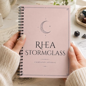May include: A light pink spiral-bound campaign journal with a black wire coil. The cover features a crescent moon graphic, the text RHEA STORMGLASS, and the words CAMPAIGN JOURNAL centered below. The design includes a thin black border frame.