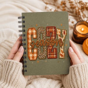 May include: An olive green spiral-bound notebook featuring the word COZY in large block letters with varied patterns including orange plaid, leopard print, and autumn leaves, with the word season written in cursive script across the centre.
