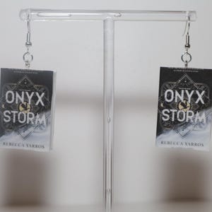 May include: A pair of earrings featuring miniature book covers. Each earring displays the title "ONYX STORM" with a geometric design and the author's name, Rebecca Yarros, against a dark background. The earrings hang from silver-colored hooks.