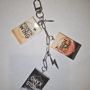 May include: A silver keychain featuring three miniature book charms: "Fourth Wing," "Iron Flame," and "Onyx Storm" by Rebecca Yarros. The keychain includes a lightning bolt charm and a chain link design.