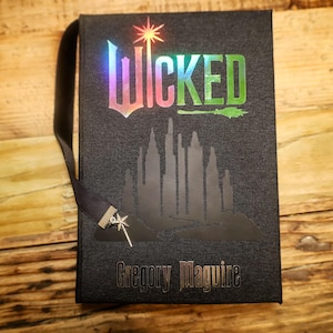 May include: A black book with the title "Wicked" in iridescent lettering and a silhouette of a city skyline. The author's name, Gregory Maguire, is printed at the bottom. A black ribbon bookmark with a star charm is attached.