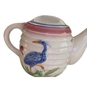 May include: A white ceramic teapot with a blue and white bird design. The teapot has a pink rim and a white handle.