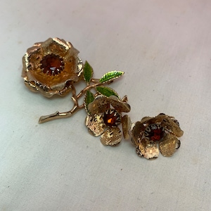 May include: A vintage gold-tone floral brooch and matching earrings set. The brooch features a large flower with a brown center stone, and the earrings have smaller matching flowers. Green and gold leaves accent the floral design.