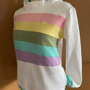 May include: White hoodie with a rainbow stripe pattern in pink, yellow, green, blue, and purple. The hoodie has a light blue hood lining.